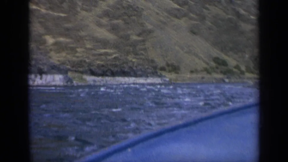 1963 Washington home-movie frame of a blue boat coasting along rough water below steep mountain terrain.