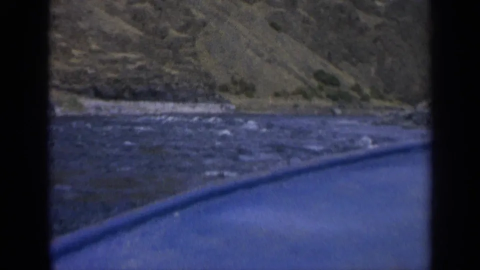 1963 home-movie frame showing a low passenger view over the boat edge toward river current and a rocky Washington hillside.