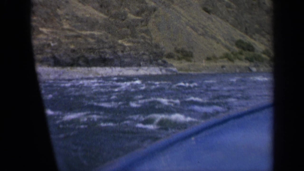 1963 Washington 8mm view from a boat as choppy blue water passes below a rugged grassy bank.