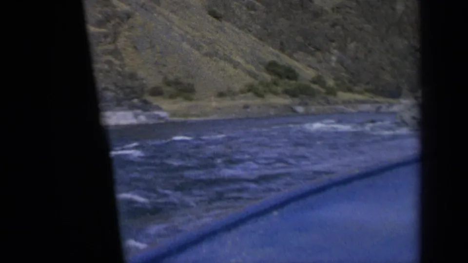 1963 Washington home-movie frame showing moving river water and a sparsely vegetated mountain slope from a car window.