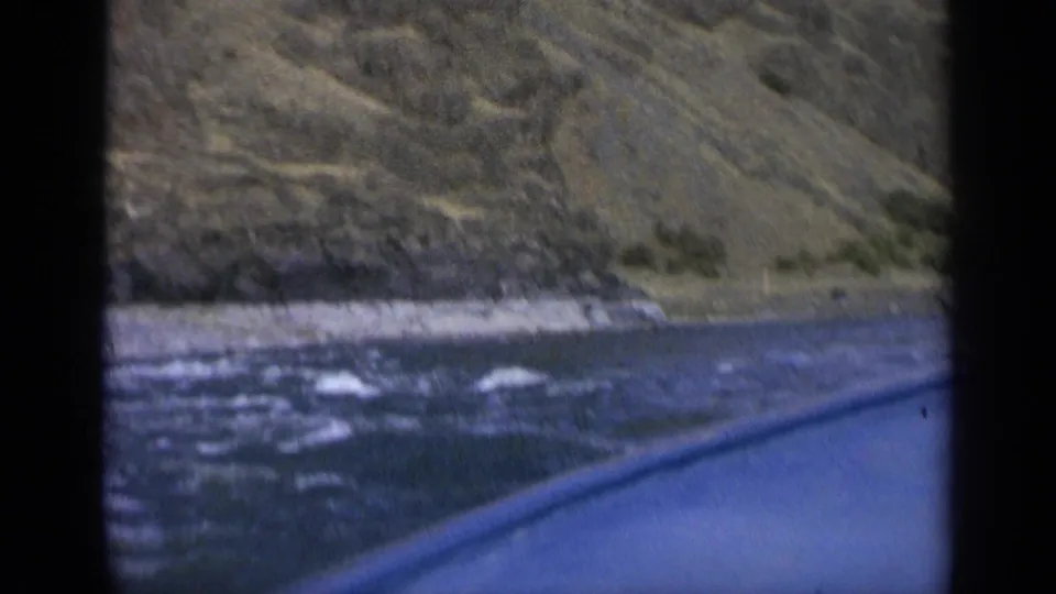 1963 Washington road-view frame of a blue-green river, whitewater, and rocky mountain terrain from a moving vehicle.
