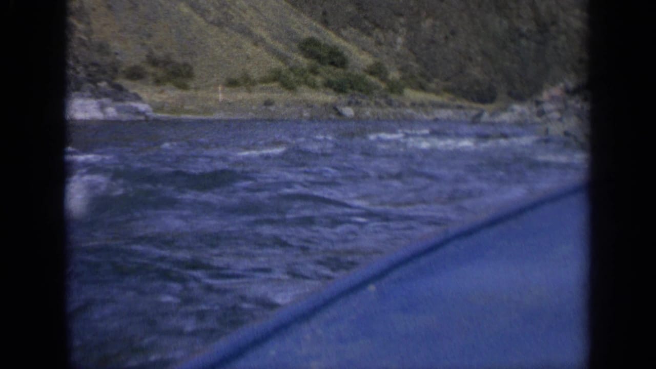 1963 home-movie frame from a boat on choppy Washington river water, with rocky slopes and sparse shrubs in the background.