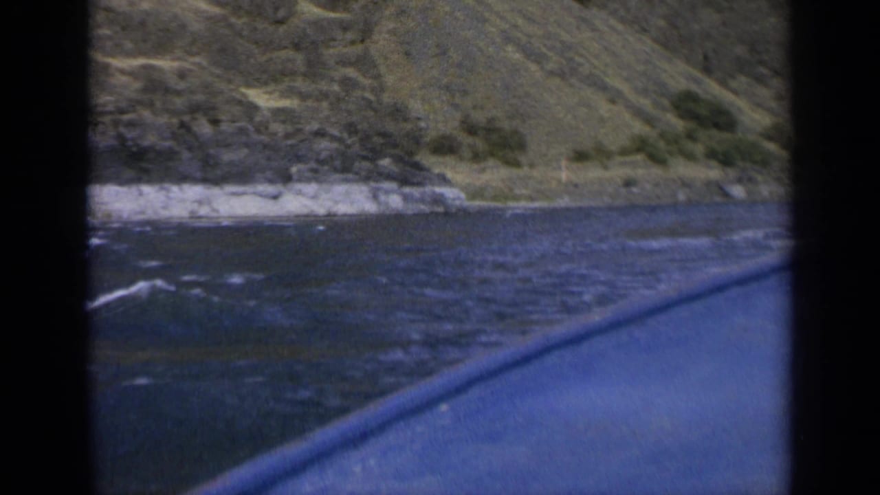 Blue boat edge cuts across dark water with a rocky bank and hillside behind it.