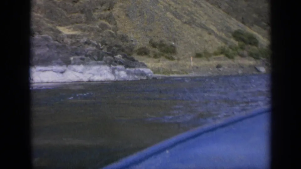 A 1963 home-movie view over a boat edge toward a rocky Washington riverbank and dry hillside.