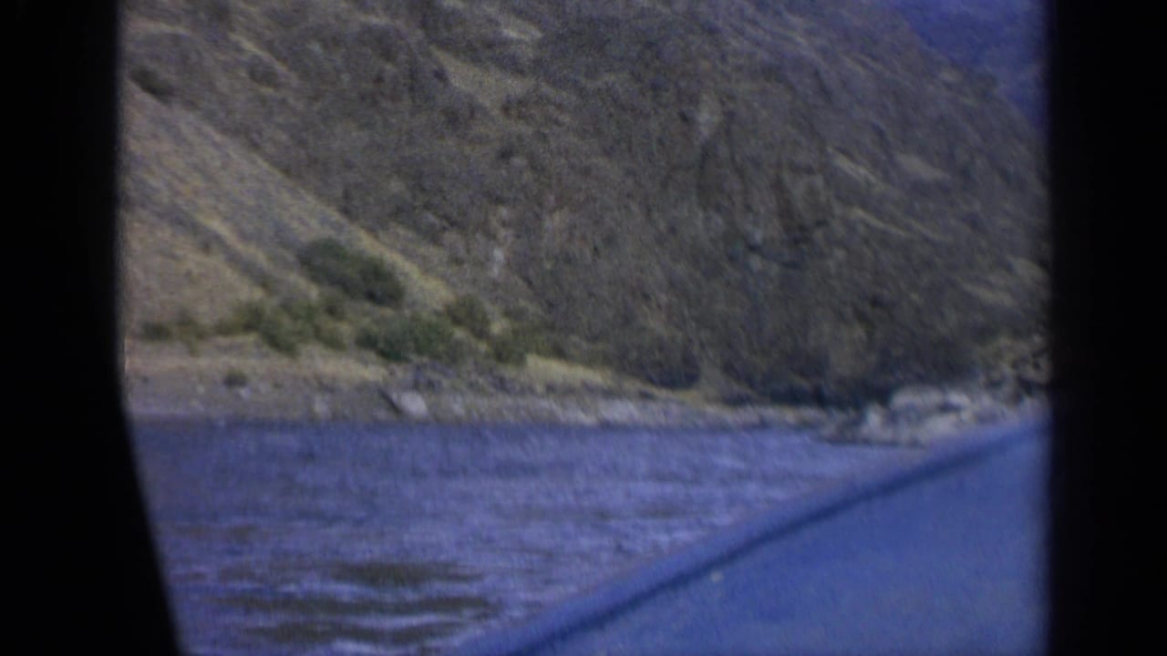 1963 Washington 8mm frame from inside a boat, looking over rippled lake water toward a rugged mountain slope.