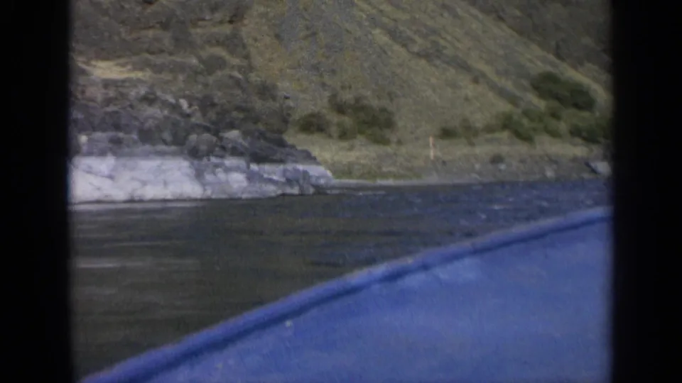 The blue edge of the boat crosses the foreground as 8mm footage looks toward a rugged Washington lakeshore.