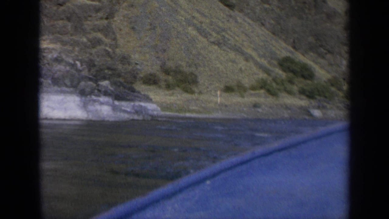 1963 Washington 8mm view from a moving boat, looking across blue lake water toward a rocky, grassy mountain shoreline.