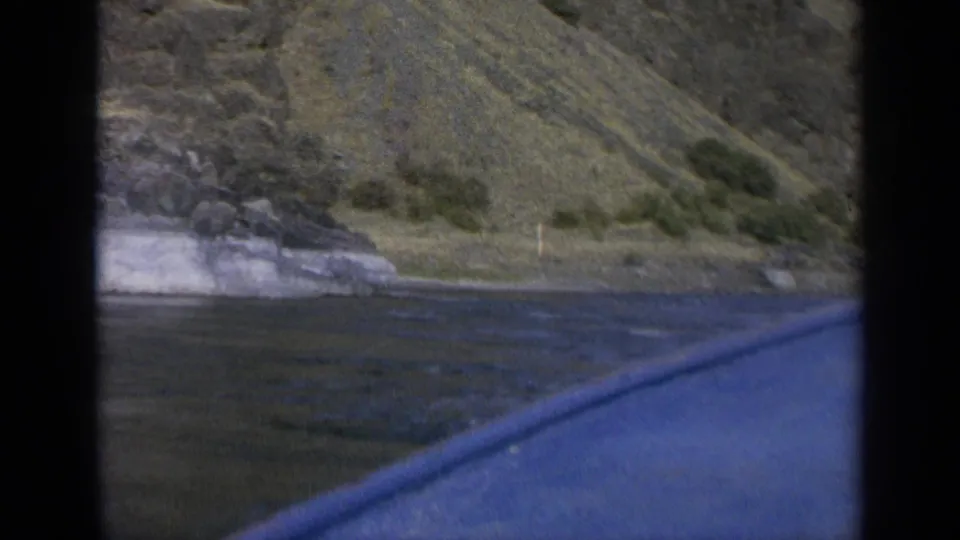 1963 Washington 8mm view from a moving boat, looking across blue lake water toward a rocky, grassy mountain shoreline.
