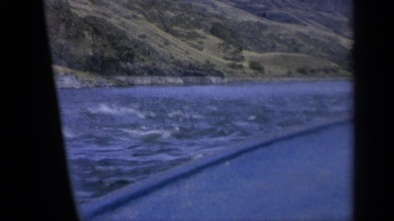 1963 home-movie frame showing lake water from a moving boat, with distant shoreline, grassy slopes, and dark window framing.