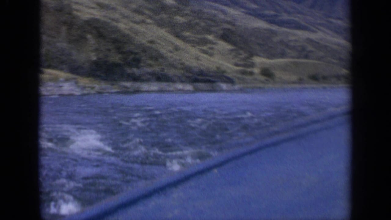 Rushing river and dry hills seen through a dark vehicle window frame.