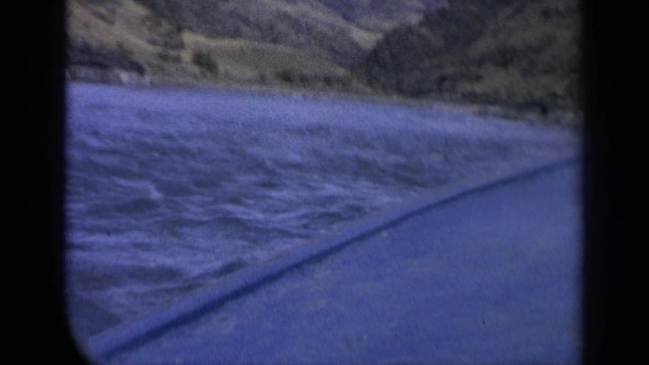 Home-movie view of blue river water and a grassy Washington bank from a moving vehicle window.