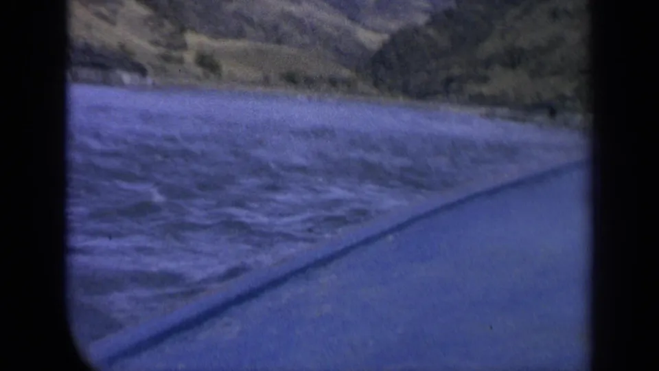 Home-movie view of blue river water and a grassy Washington bank from a moving vehicle window.