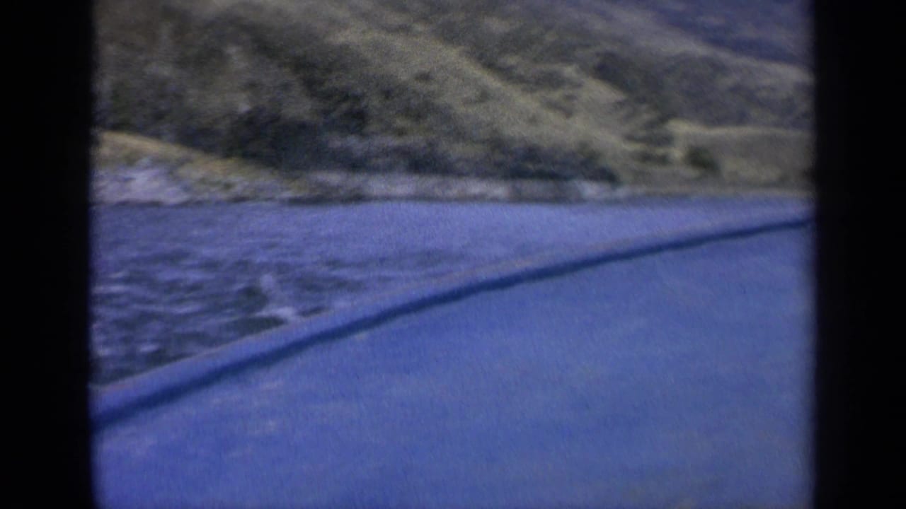 1963 Washington 8mm frame showing blue lake water, a sloping green shoreline, and a paved edge in the foreground.