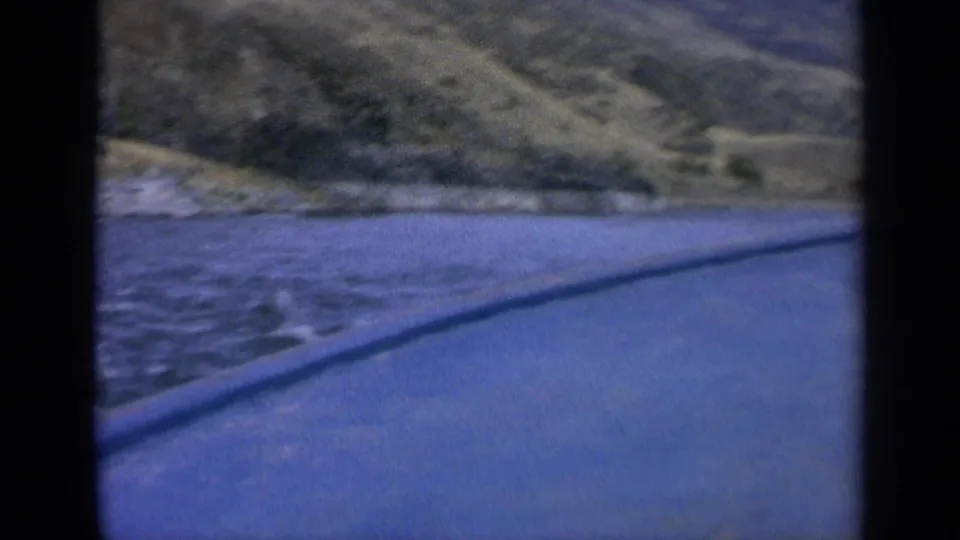 1963 Washington 8mm frame showing blue lake water, a sloping green shoreline, and a paved edge in the foreground.