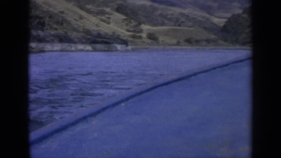 1963 Washington 8mm frame showing lake water, a low shoreline, green hills, and a diagonal paved or vehicle-side edge.