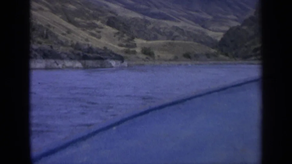 1963 Washington 8mm view across blue river water toward a dry hillside and low shoreline vegetation.