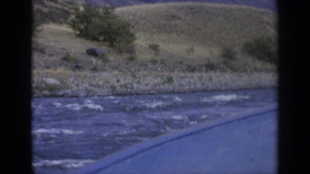 1963 8mm Washington river view from a moving boat, with water, rocky bank, dry rolling hills, and sparse vegetation.