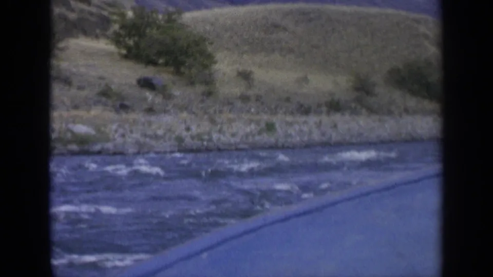 1963 8mm Washington river view from a moving boat, with water, rocky bank, dry rolling hills, and sparse vegetation.