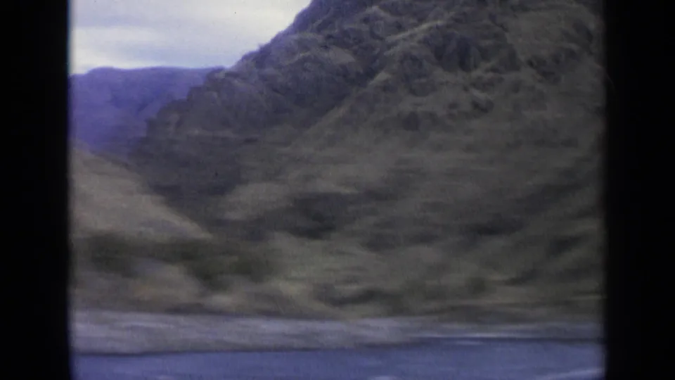 1963 8mm Washington river landscape with canyon-like slopes, foreground water, sparse vegetation, and a muted sky.