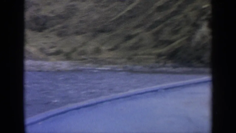 1963 Washington 8mm view from a moving car, looking past a road edge toward river water and steep mountain terrain.