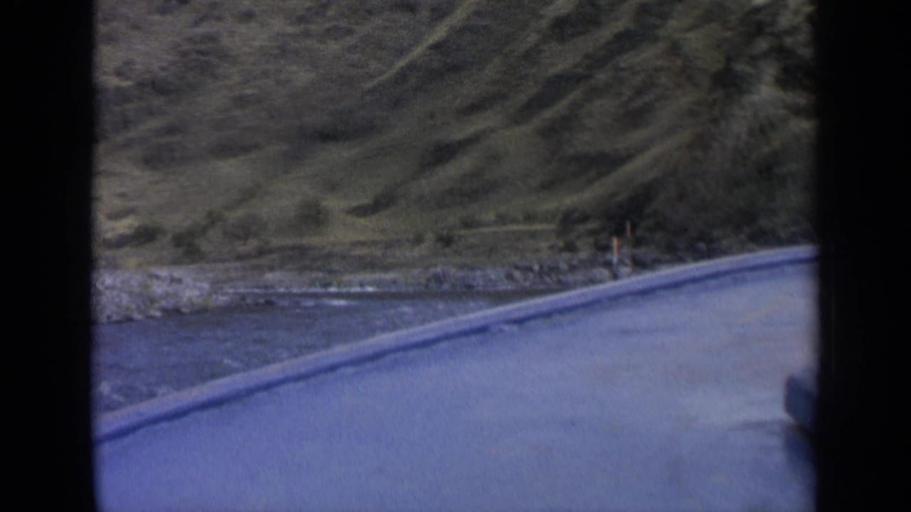 Washington road-trip frame with paved edge, riverbank, hillside, and small distant roadside figures or posts not clearly identifiable.