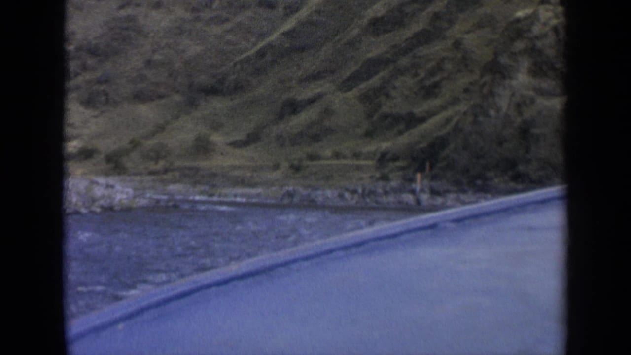 1963 Washington 8mm frame showing a river, roadside edge, rocky bank, and steep green hillside viewed from a car window.