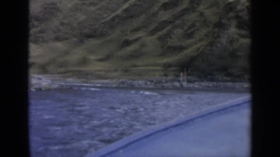 1963 Washington roadside view with river water, a paved road edge, rocky bank, trees, and a distant person near the water.