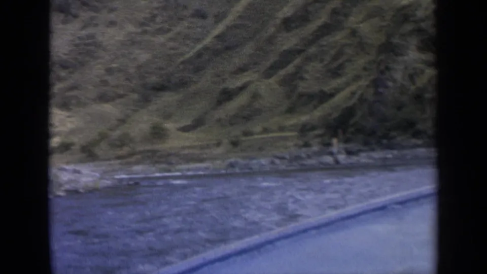 1963 Washington 8mm view across a river from a moving car, with the road edge, rocky bank, hillside, and trees in frame.