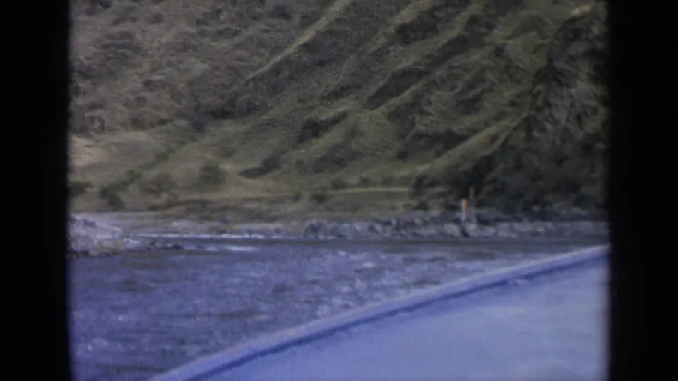 1963 Washington frame showing a river beside a paved road, with a rocky bank, hillside, conifers, and a distant person near shore.