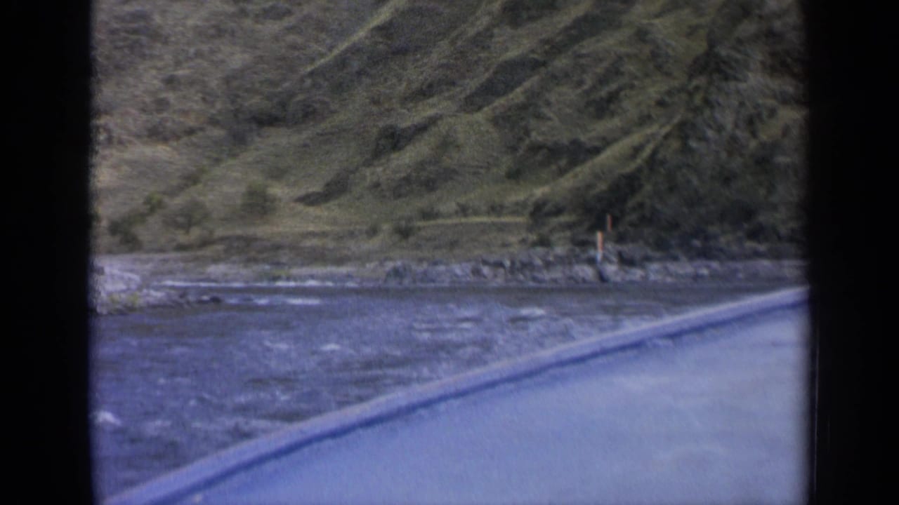 1963 Washington 8mm frame showing rippling water, rocky shore, a pale road or boat edge, and steep green hills beyond.
