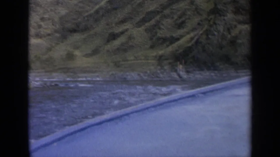 1963 Washington shoreline view from a moving boat, with blue rippling water, rocks, and steep grassy terrain in the background.