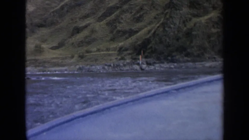 Vintage 8mm Washington travel frame with rippling water, rocks, a small shore marker, and steep green hillside beyond.