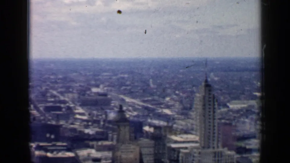 1957 Chicago downtown appears in a soft 8mm aerial view, with a tall tower, low rooftops, and a distant horizon beyond the city.