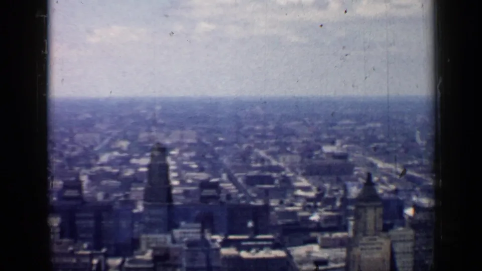 1957 Chicago skyline seen from an elevated window, showing clustered towers, rooftops, and a pale horizon under aged 8mm film grain.
