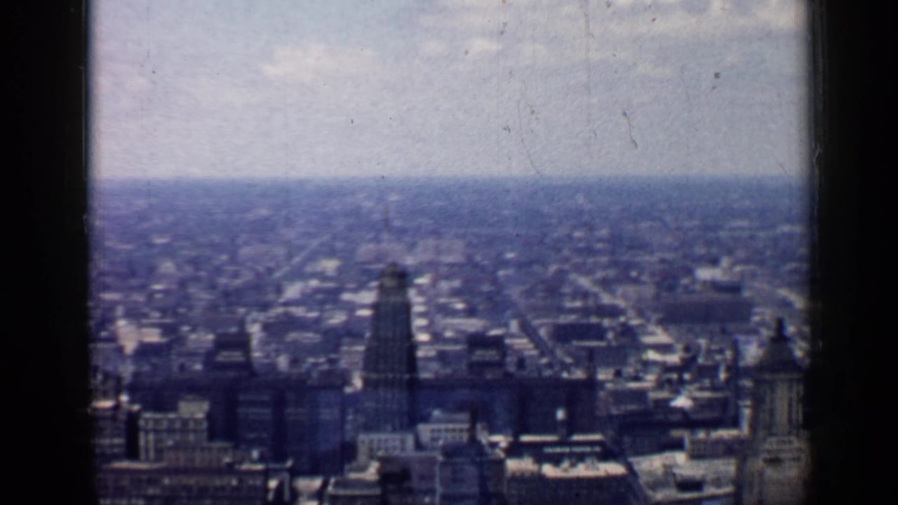 1957 Chicago skyline from a high vantage point, with distant towers, low city blocks, hazy horizon, and dark film or window edges.