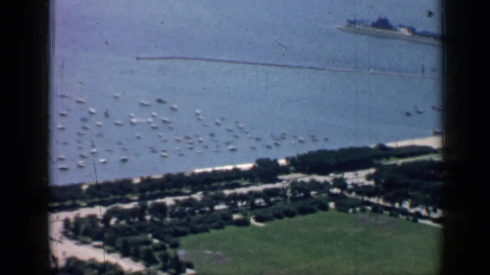 Aerial 8mm view over Chicago waterfront, with anchored boats, a tree-lined shore, open lawn, and a distant pier or breakwater.