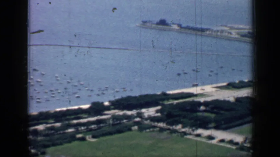 1957 Chicago shoreline frame with small boats offshore, a pale roadway along the water, green parkland, trees, and a long pier in the distance.