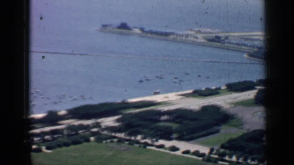 Chicago 1957 8mm frame looking down on a waterfront lawn and shoreline road, with scattered boats and a long pier or breakwater beyond.
