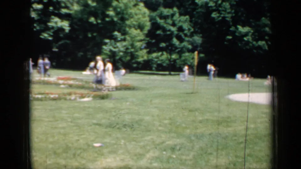 In a Chicago park scene from the 1957 home movie, women in pale dresses walk across open grass near low flower beds.