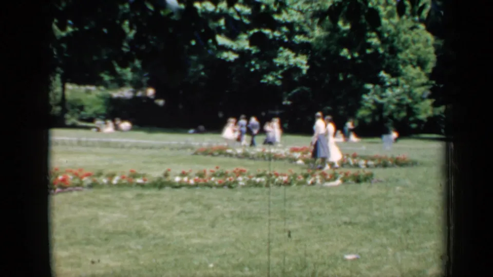 Women and other distant visitors move through a landscaped Chicago park area with flower beds, grass, and leafy trees.