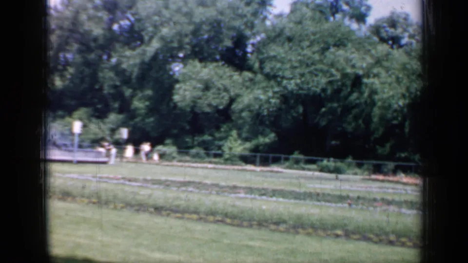 1957 Chicago 8mm frame with garden rows in front and small distant figures near signs along a tree-lined path.
