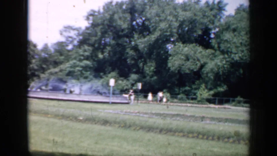 1957 Chicago home-movie frame showing sprinkler water over lawn, garden beds, distant walkers, signs, and mature trees.