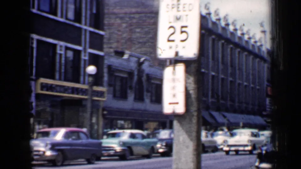 1957 Chicago street view with classic cars passing brick storefronts beneath a prominent 25 mph speed limit sign.