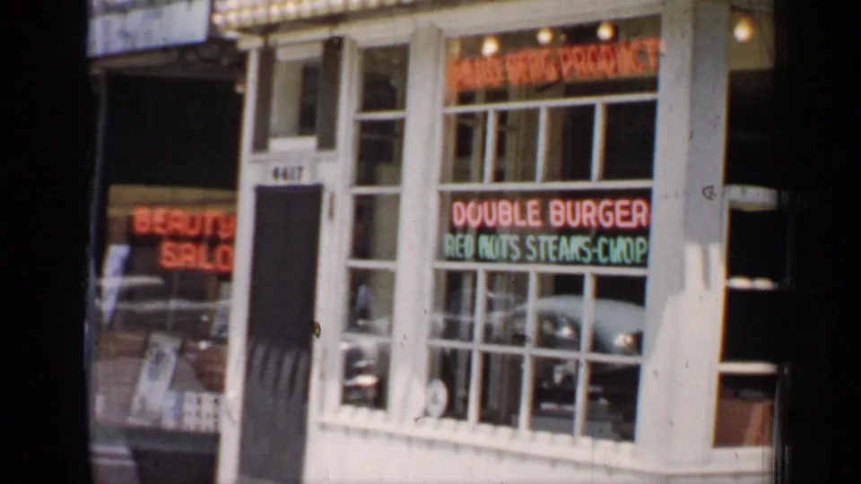 1957 Chicago 8mm frame showing the shop entrance, marquee edge, and neon window text advertising burgers and other food.