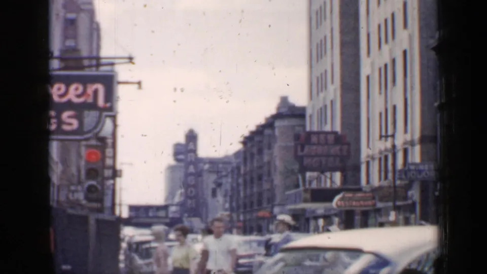 1957 Chicago downtown street scene with pedestrians crossing near mid-century cars, tall buildings, and visible commercial signs.