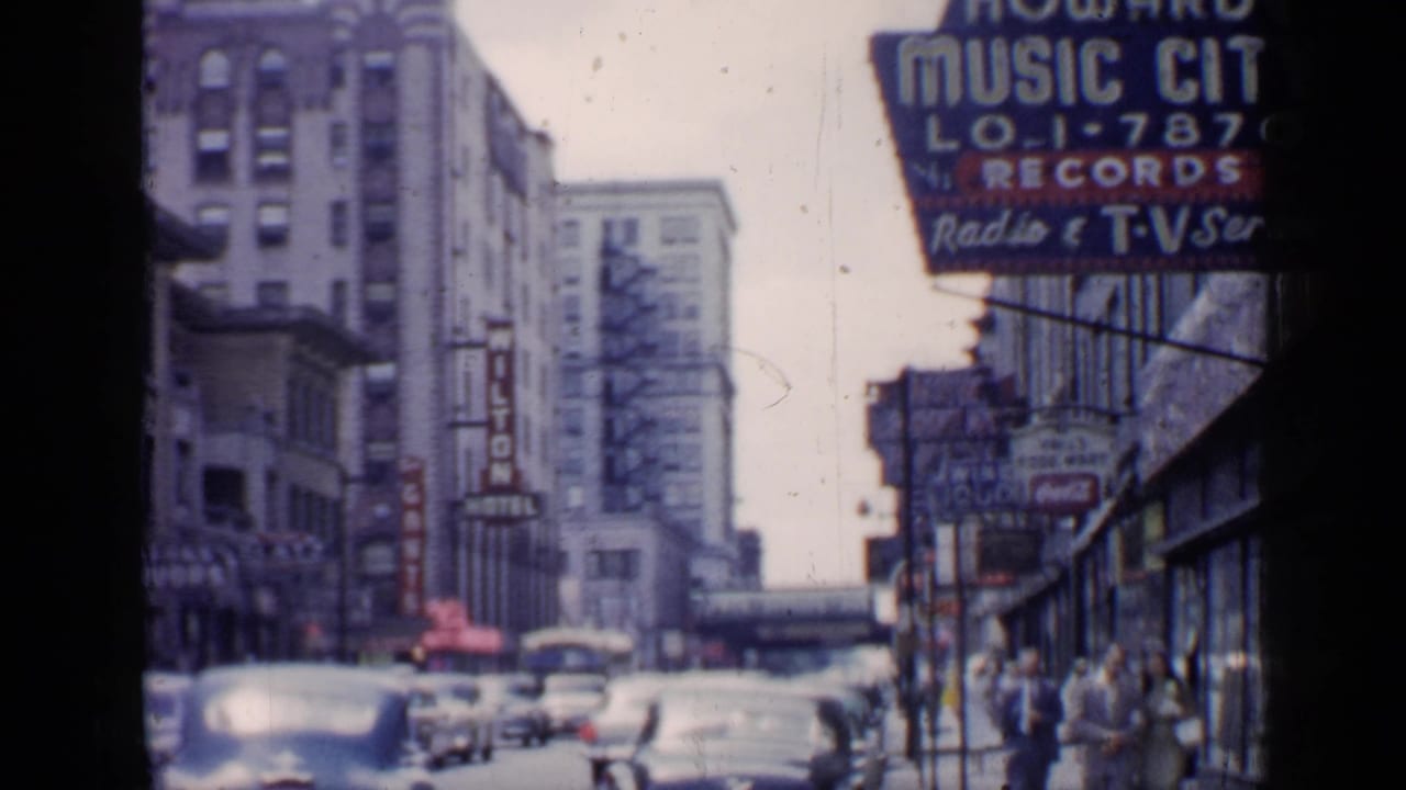 1957 Chicago street view with the Wilton Hotel sign, Howard Music City storefront sign, vintage cars, and pedestrians along the sidewalk.