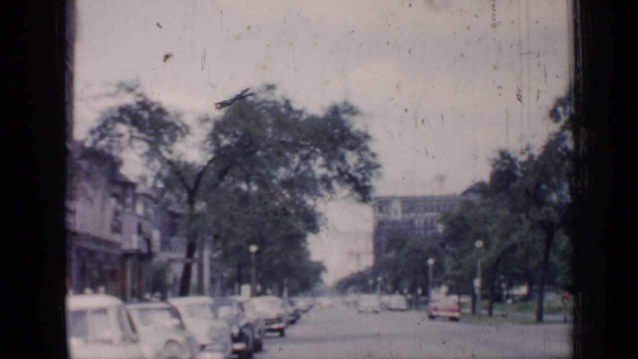 1957 Chicago street view with curbside automobiles, leafy trees, streetlamps, and a distant building seen through scratched 8mm film.