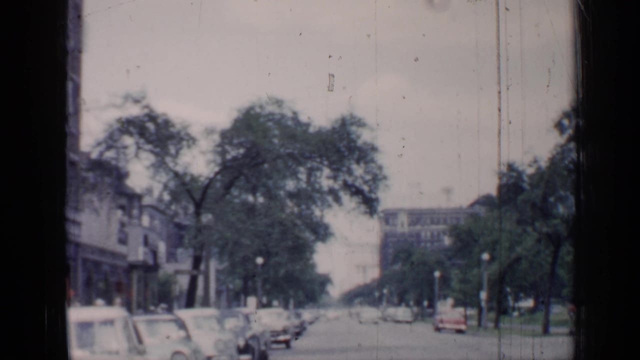 1957 Chicago home-movie frame showing parked automobiles along a broad street with trees, streetlights, and urban buildings beyond.