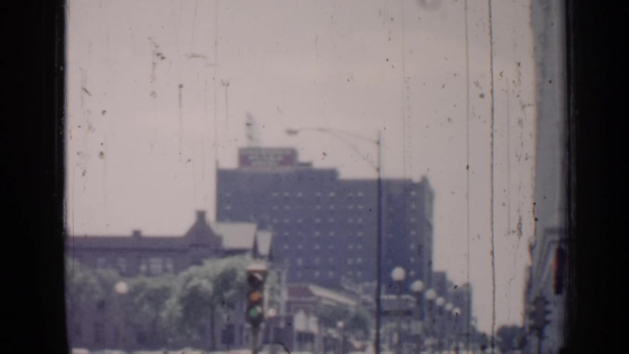 1957 Chicago 8mm view shifts toward a street corner with a traffic signal, globe streetlights, trees, and a taller building in the background.