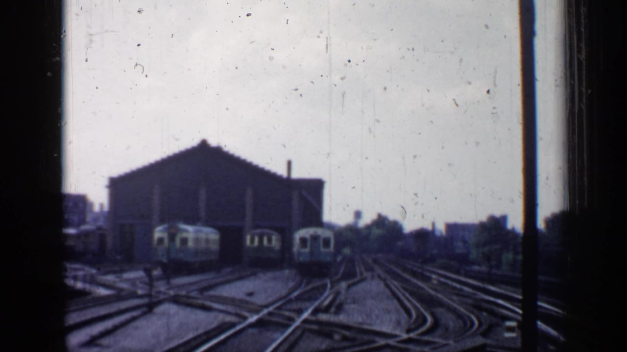 Rail yard tracks with several pale train cars near a large shed under an overcast sky.